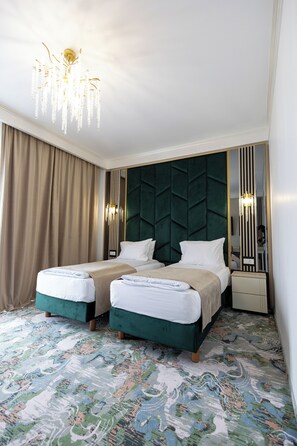 Ein Hotelzimmer mit zwei Betten, einer Kristallleuchtre, einem grünen Stoffpolster-Kopfteil und einem gemusterten Teppich.