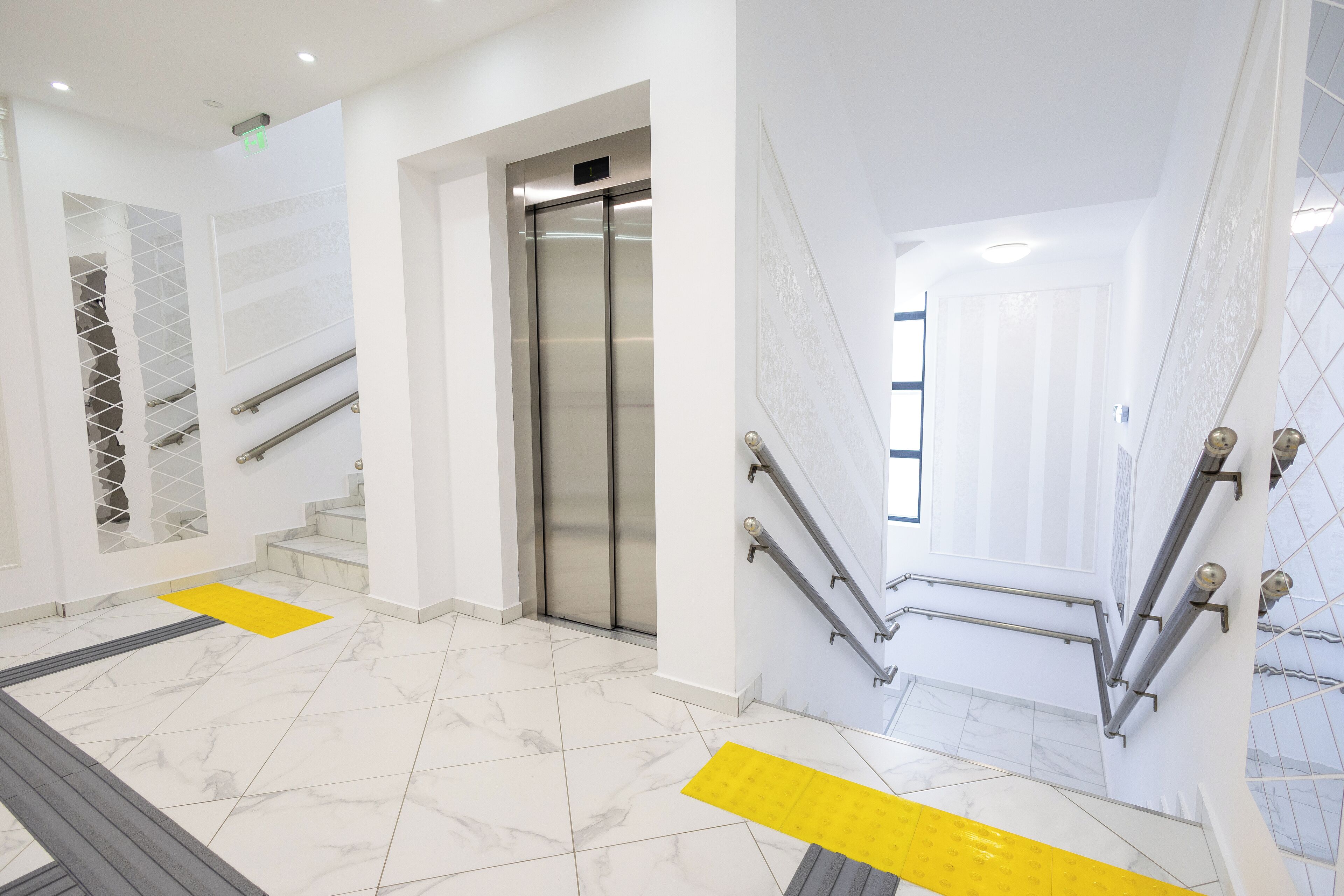 A modern hotel lobby with marble flooring, a central elevator, and a staircase with handrails.