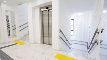Un moderno vestíbulo de hotel con suelo de mármol, un ascensor central y una escalera con barandillas.