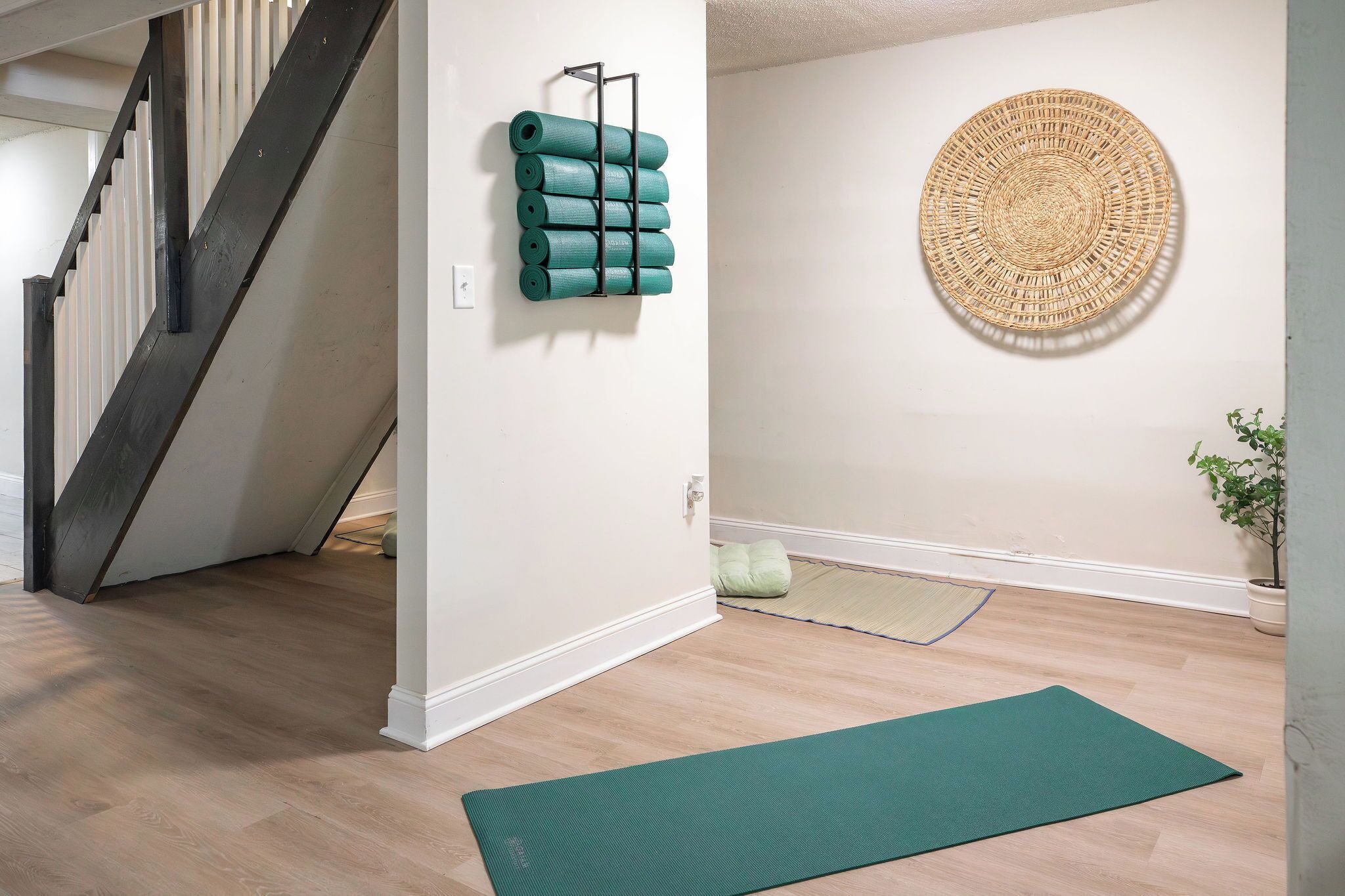 Um quarto com escada de madeira, suporte de yoga fixado na parede, um grande painel de tear na parede, um tapete de yoga verde no chão e um vaso com planta.