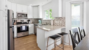 A modern kitchen with white cabinets, stainless steel appliances, a granite countertop, and a small island with bar stools. - Mini Golf | FirePit | GameRoom | 4BRs | Cedar Park (Cedar Park)