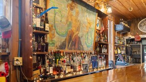 Uma área de bar com teto de madeira, um mural e prateleiras repletas de garrafas de bebidas diversas.