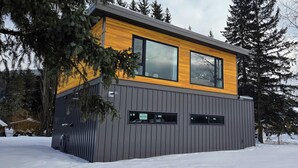 En modern byggnad med en övre del i trä och en grå nedre del, omgiven av snö och träd.