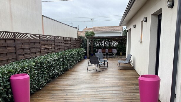 A wooden deck with a low wall and a hedge, two chairs, and a table under a covered area.