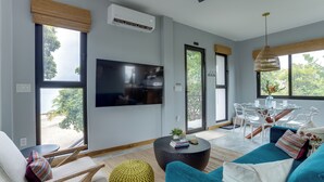A modern living room with a flat-screen TV, a dining area with a glass table, and large windows offering a view of the outdoors. - Take Two! Private Beach House! (Placencia Village)