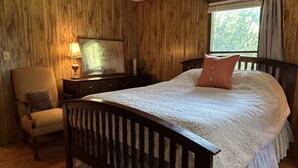 A bedroom with a wooden paneled wall, a bed with a white bedspread and a pink pillow, a chair with a patterned cushion, a nightstand with a lamp, and a window with a view of trees. - Retro Cottage Farmstay
Mid-term Comfort for Family Relocations/Professionals (Newton)