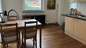 A kitchen with wooden cabinets, a dining table with chairs, a radiator, and a window with a view of a building outside. - Centrally located, small apartment with kitchen and shower room 2 (Hamburg)