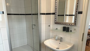 A bathroom with a glass-enclosed shower, a white sink with a mirror above it, and a black-tiled floor. - Centrally located, small apartment with kitchen and shower room 2 (Hamburg)