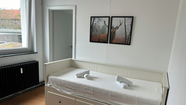 A bedroom with a white bed, a radiator, a window, and two framed pictures on the wall. - Centrally located, small apartment with kitchen and shower room 2 (Hamburg)
