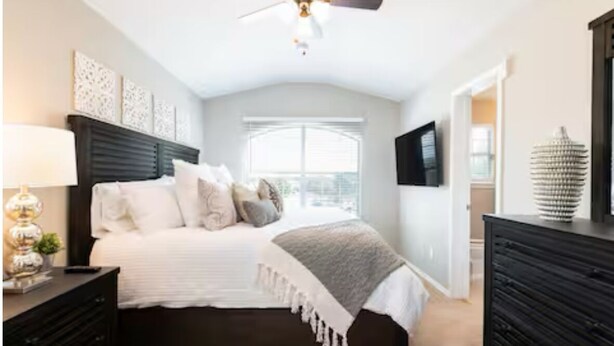 Une chambre avec un lit en bois foncé, du linge de lit blanc, un ventilateur de plafond et une grande fenêtre.