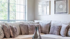 Un salon avec un canapé beige, des coussins à motifs, un vase en verre posé sur une table sombre, et deux œuvres d’art encadrées accrochées au mur.