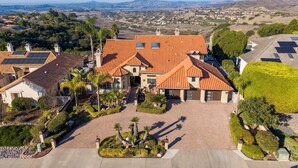 A large, single-story house with a red-tiled roof, surrounded by a well-maintained garden with palm trees and a paved driveway. - Scenic Luxury Oasis: Views, Pool, Spa, Game & Movie Rms (Bonita)