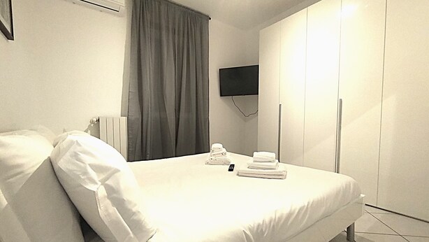 A hotel room with a bed, pillows, a TV, a wardrobe, and a set of towels. - HH Monferrato (Alessandria)
