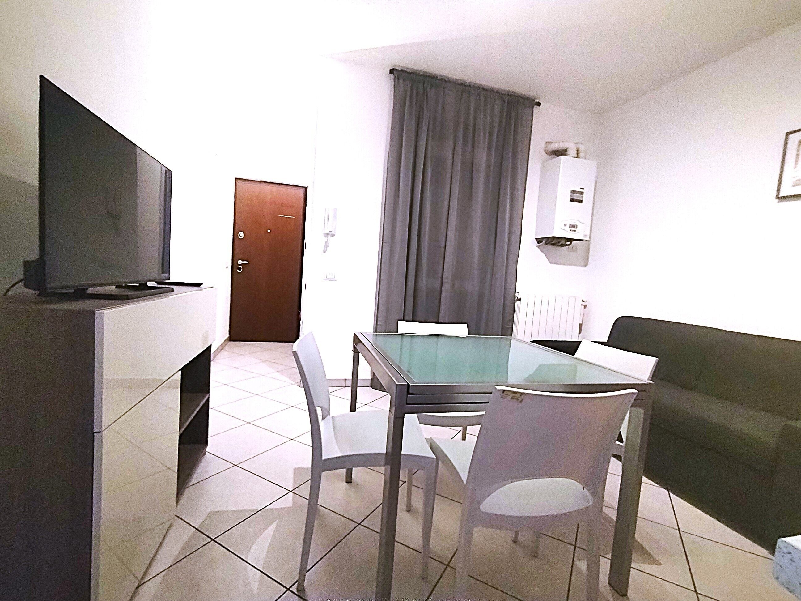 Una stanza piccola e ben illuminata, con televisione su un mobile, un tavolo da pranzo con sedie, un divano, una porta e una finestra con tende.