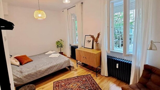 A bedroom with a bed, a wooden cabinet, a radiator, a window with curtains, and a hanging light fixture. - KK30 Apartment (Budapest)