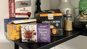 A shelf with various packaged food items including a box of Segafredo Zanetti INTERMEZZO, a box of LUNGO CREMA, a bottle of Burla ETELE, a box of Moris, and a box of Herbaria Winter Magic Tea. - KK30 Apartment (Budapest)