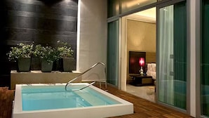Una piscina interior moderna con un deck de madera, rodeada de plantas en macetas y una puerta de cristal que da a una habitación con cama y mesita de noche.