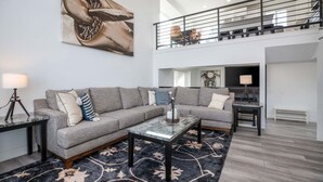 A spacious living room with a grey sectional sofa, a coffee table, and a large abstract painting. A staircase leads to an upper level with a dining area. - Cottonwood Basecamp-Game Room, 6TVS, Near Canyons! (Sandy)