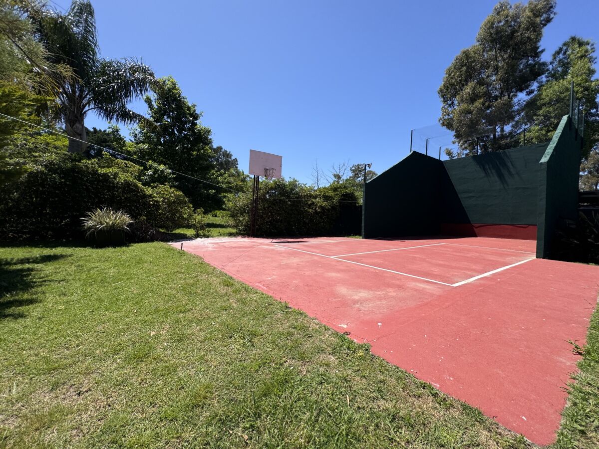An outdoor tennis court with a red surface, surrounded by grass and trees.