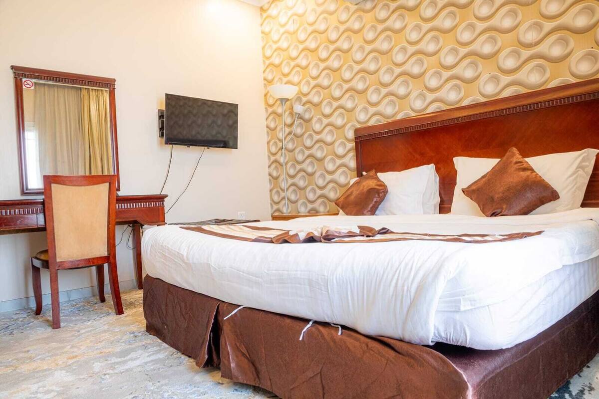 A hotel room with a large bed, a desk with a chair, a television, and a patterned wall.