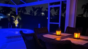 Un área de comedor al aire libre con sillas y mesas de mimbre, iluminada por faroles, con vistas a una piscina y palmeras.