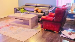 A living room with a sofa, a red chair, a coffee table, and a window with blinds.