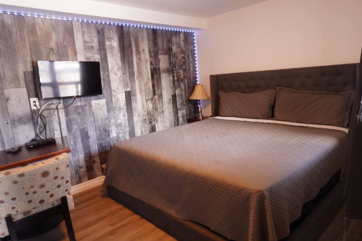 A bedroom with a wooden wall, a flat-screen TV mounted above a desk, a bed with a brown headboard, and a lamp on a bedside table.