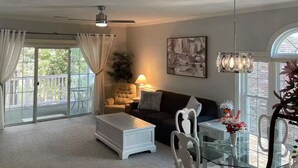 Ein geräumiges Wohnzimmer mit Deckenventilator, einem Glastisch mit Stühlen, einer Couch und einem großen Fenster mit Vorhängen.