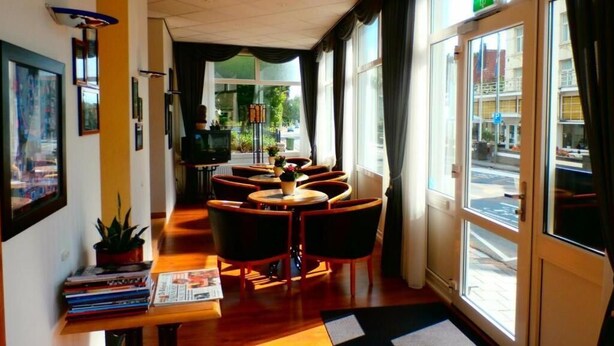 Un hall d’hôtel avec un comptoir d’accueil en bois, plusieurs tables rondes et des chaises. Les murs sont ornés de tableaux encadrés et un pot de fleurs est posé sur le comptoir.
