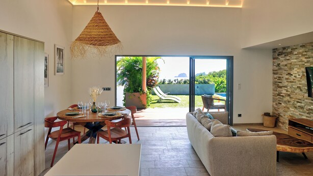 Ein modernes Wohnzimmer mit einer Couch, einem Esstisch und Stühlen. Ein großes Fenster bietet Blick auf Grünflächen und einen Pool.