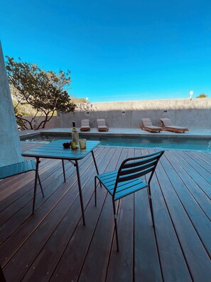 Una terrassa de fusta amb piscina, gandules i una taula amb una ampolla i vasos.
