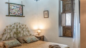 A bed with a patterned bedspread, a decorative headboard, a bedside lamp, a map pillow, a framed picture on the wall, and a window with a wooden shutter.