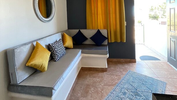 Een moderne woonkamer met een grijze bank, gele en blauwe kussens, een blauw gestreept vloerkleed, een plafondventilator en een groot raam met gele gordijnen.