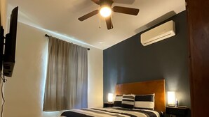 Een hotelkamer met een bed, een plafondventilator, een airconditioningapparaat, een raam met gordijnen en een aan de muur bevestigde televisie.