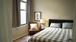 A bedroom with a checkered bedspread, a chair with a cushion, a nightstand with a lamp, and a framed picture on the wall.