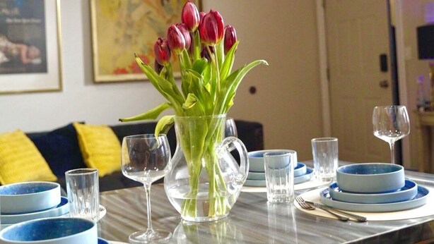 A dining table set with blue plates, a clear pitcher with red tulips, and a framed poster on the wall.