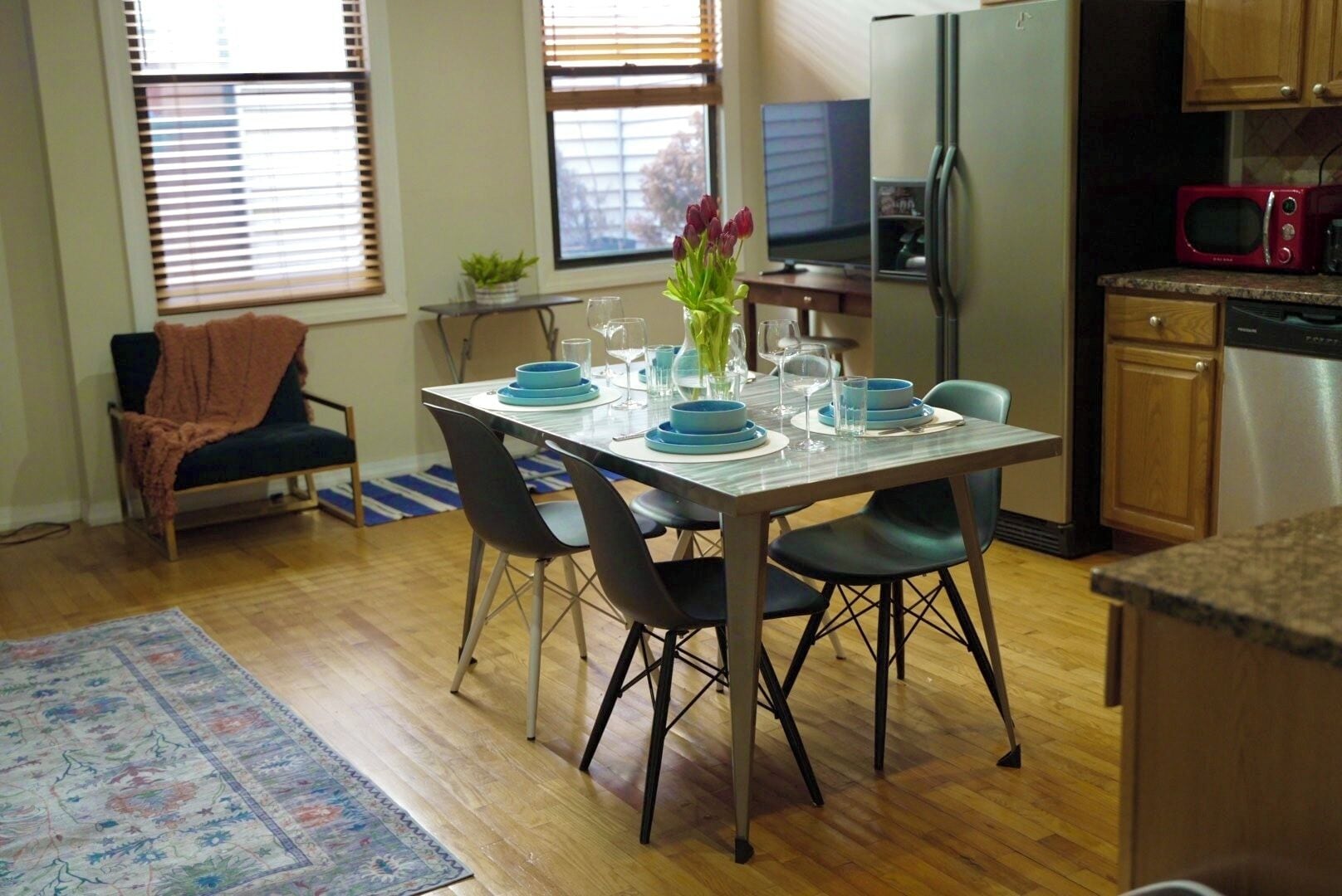 A dining area with a table set for four, surrounded by chairs. A television is mounted on the wall, and a refrigerator is visible in the background.