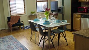 A dining area with a table set for four, surrounded by chairs. A television is mounted on the wall, and a refrigerator is visible in the background.