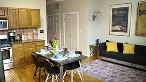 A modern kitchen with wooden cabinets, a dining table set for four, and a living area with a sofa and a rug.