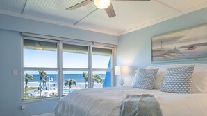 Une chambre avec un grand lit, un ventilateur de plafond et une vue sur la plage grâce à de grandes fenêtres.