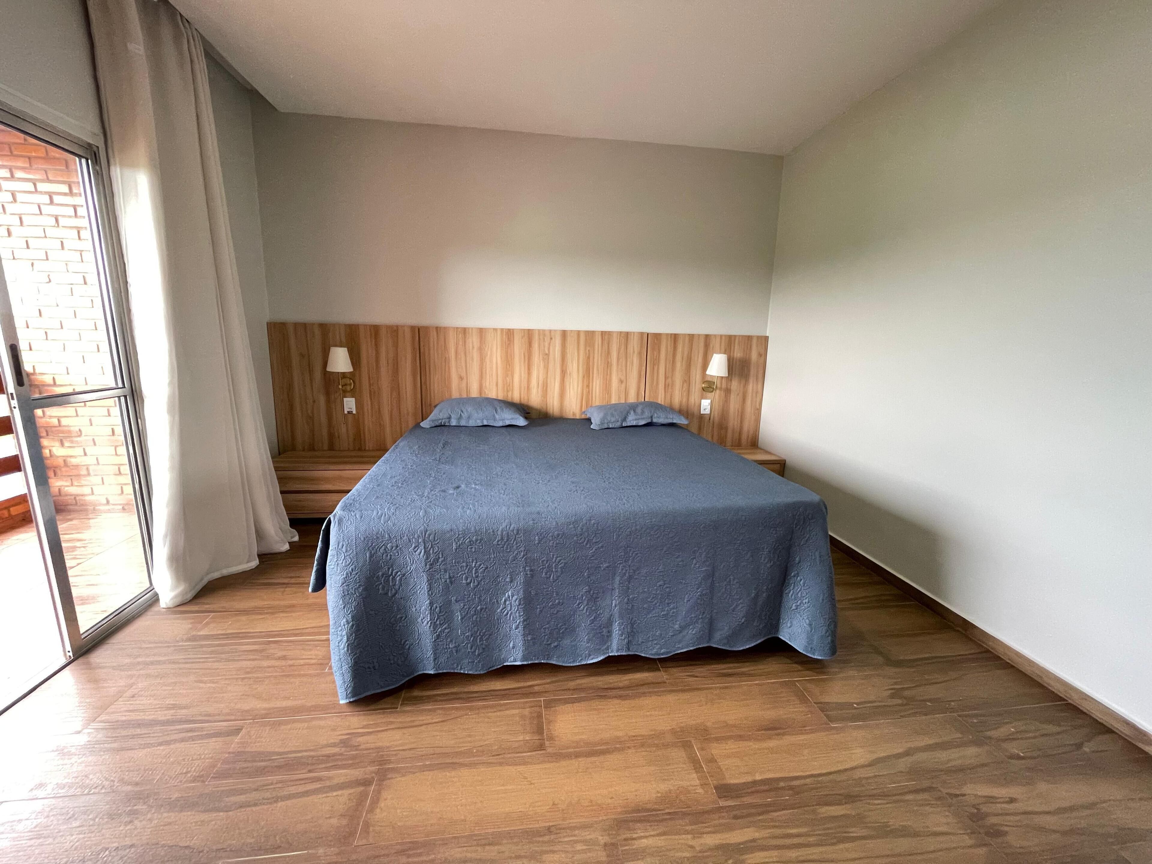 Een slaapkamer met een houten hoofdbord, een groot bed met een blauwe sprei, twee bedlampen, en uitzicht op een bakstenen muur door een schuifdeur.