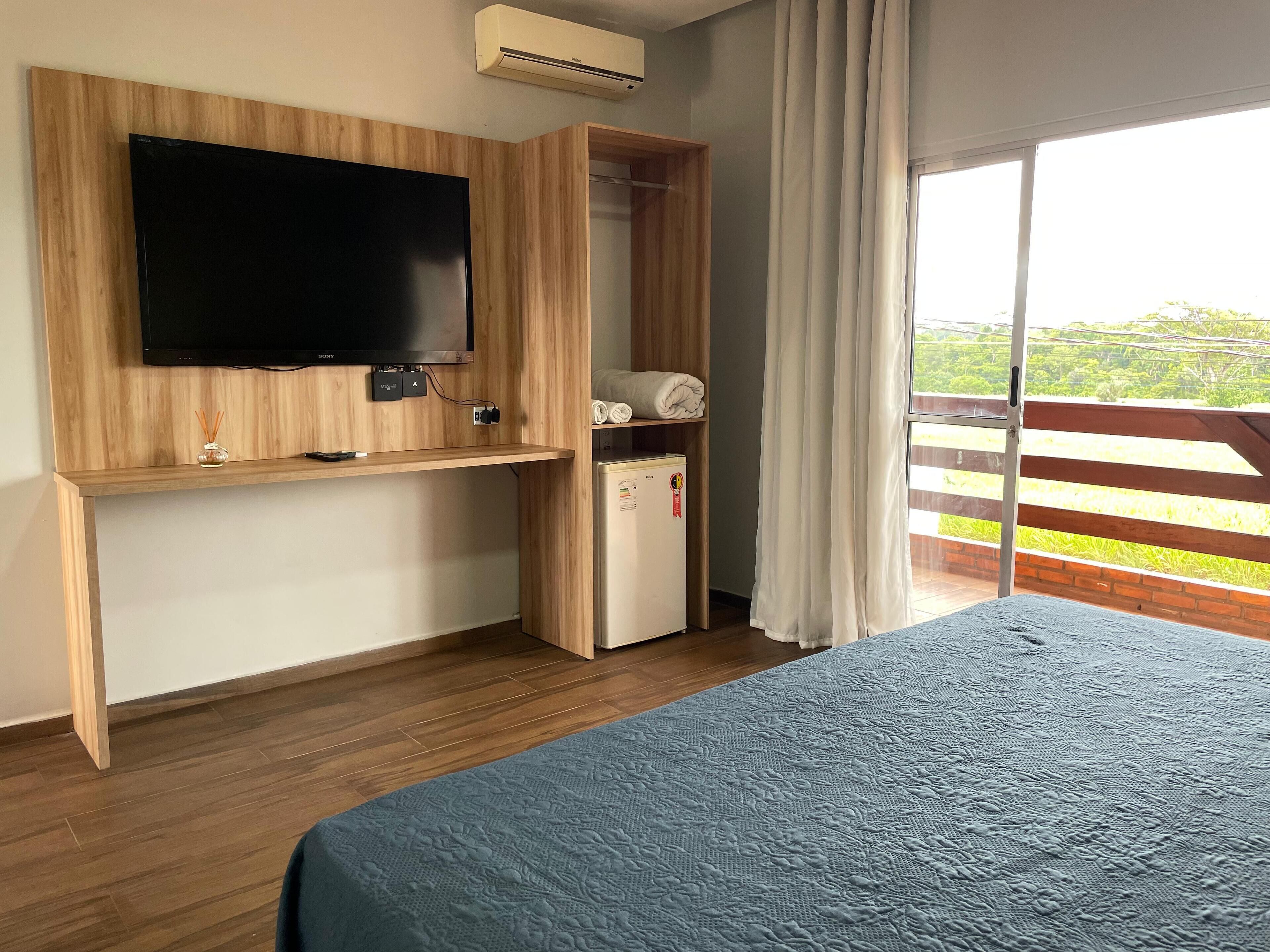 A bedroom with a large bed, a TV mounted on a wooden panel, a small fridge, and a sliding glass door to a balcony.