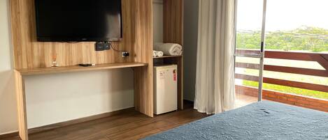 A bedroom with a large bed, a TV mounted on a wooden panel, a small fridge, and a sliding glass door to a balcony.