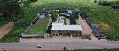 An aerial view of a building complex with multiple structures, including a main building with a large roof, adjacent smaller buildings, and solar panels. A road runs in front of the complex.