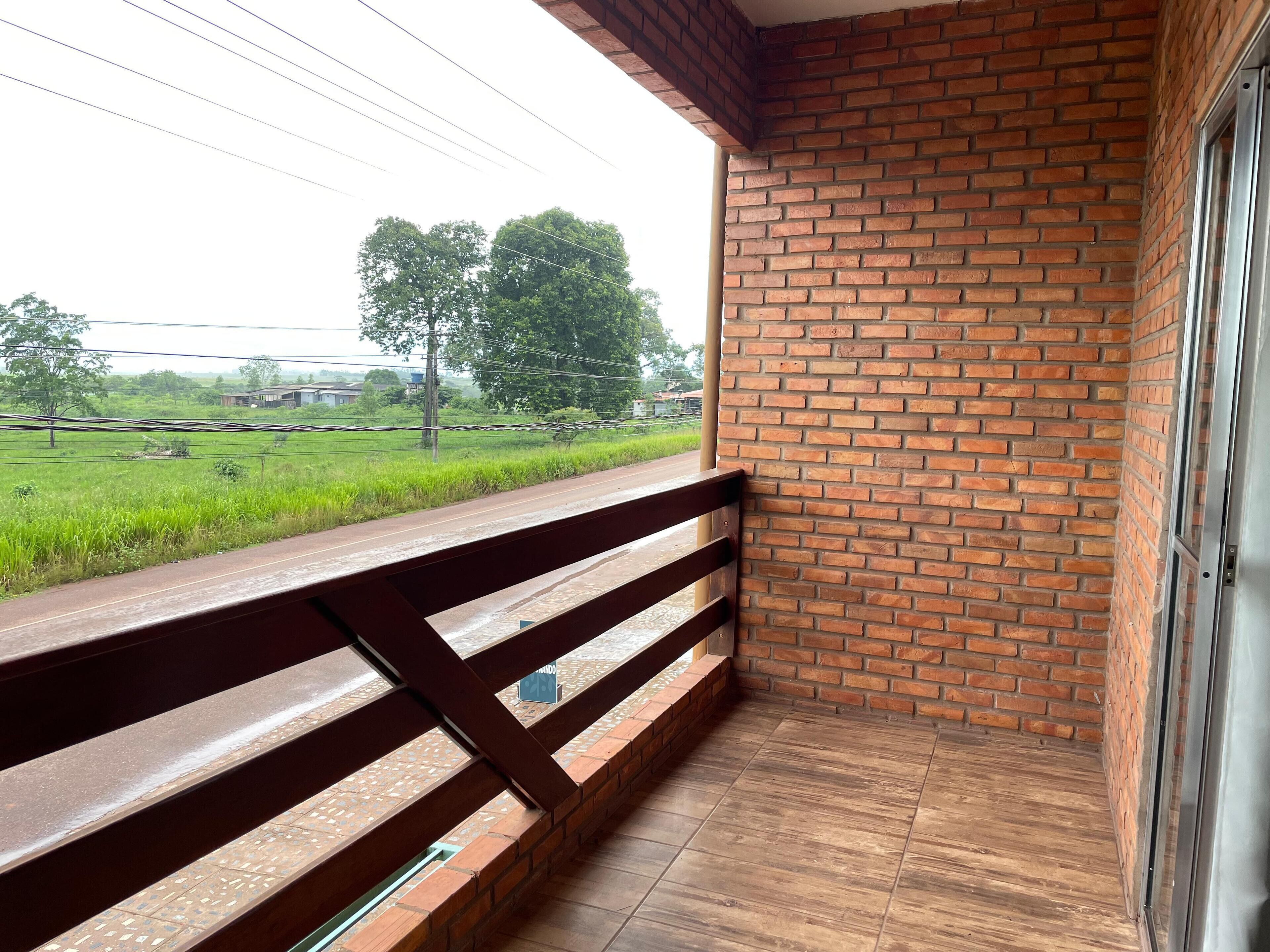 A balcony with a wooden railing overlooking a brick wall and a view of a green field with trees.