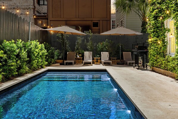 A swimming pool with lounge chairs and umbrellas, surrounded by greenery and a wooden fence.