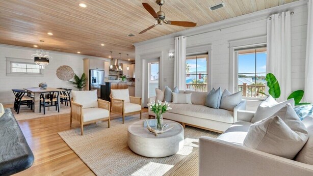 A spacious living room with a wooden ceiling, a ceiling fan, a large sofa, a coffee table, and a dining area with chairs and a table.