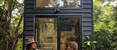 Una pequeña construcción de madera oscura, con puerta y ventanas de vidrio, ubicada sobre una terraza de madera rodeada de follaje.
