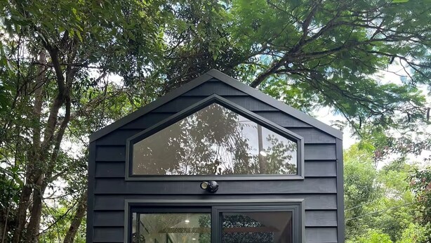 Une petite structure en bois de couleur sombre, dotée d’une porte et de fenêtres en verre, d’un banc, d’une chaise et d’une statue d’éléphant décorative, le tout installé sur une terrasse en bois.