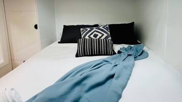 Una habitación con tragaluces, una cama con ropa de cama blanca, almohadas negras y blancas, y una colcha azul.